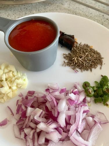 A cup of tomato puree next to chopped onions, green chillies, garlic, cumin and broken dry red chilli.