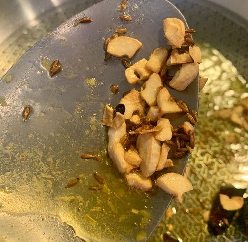 Tempered chopped garlic and cumin seeds on steel ladle. In the background spices being tempered in a pan. 