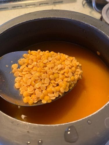 Uncooked yellow lentils in a steel ladle over a pressure cooker with water.
