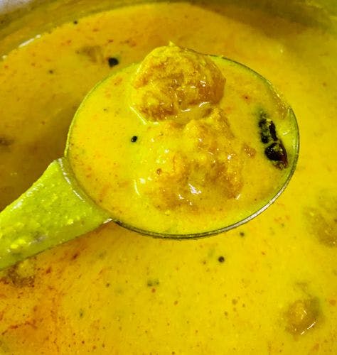 Kadhi and cooked fritters in a ladle over a pot of kadhi. 