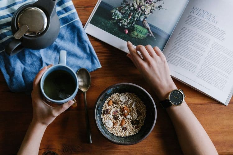 A bowl of quinoa with some dry fruits placed on a table as a person works on the table while eating from the bowl and sipping his drink from a mug.