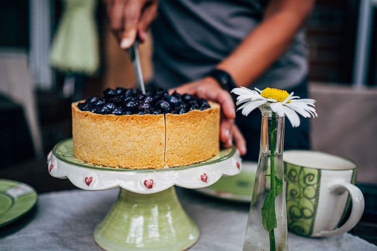 Person slicing a homemade blueberry cake on a decorated cake stand for a birthday celebration