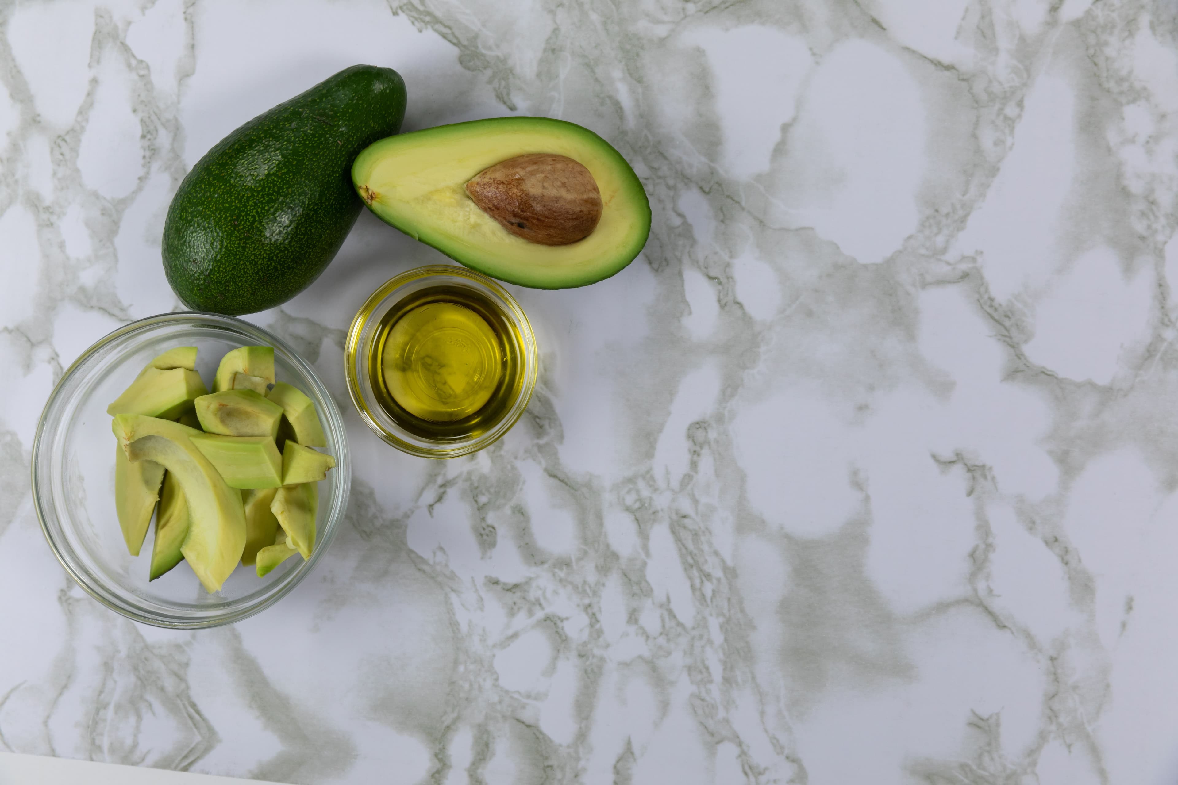 Top view of fresh avocado halves and chunks in bowls with oil on a marble surface, ideal for a healthy snack.