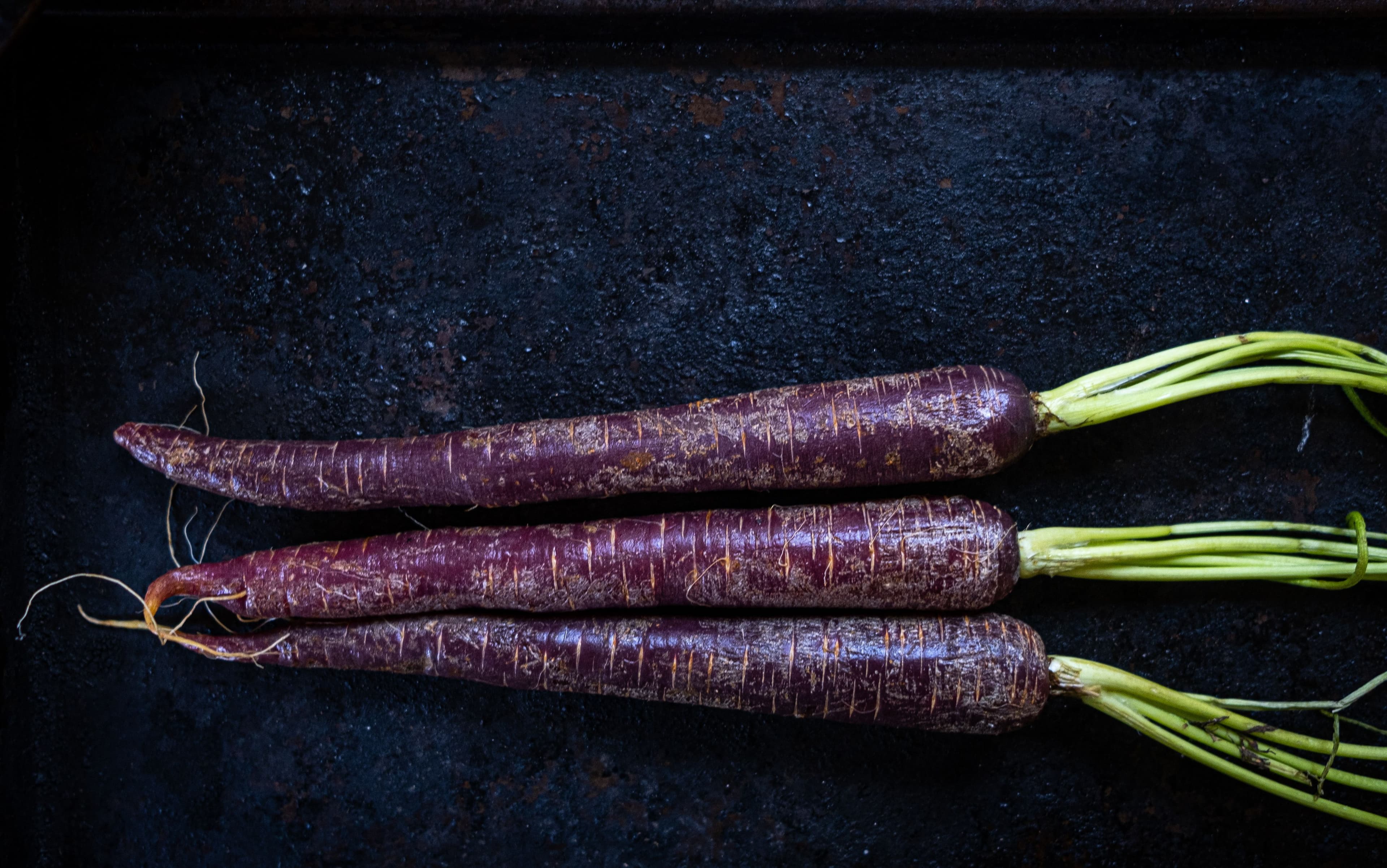Three whole black carrots with green tops on a dark rustic surface