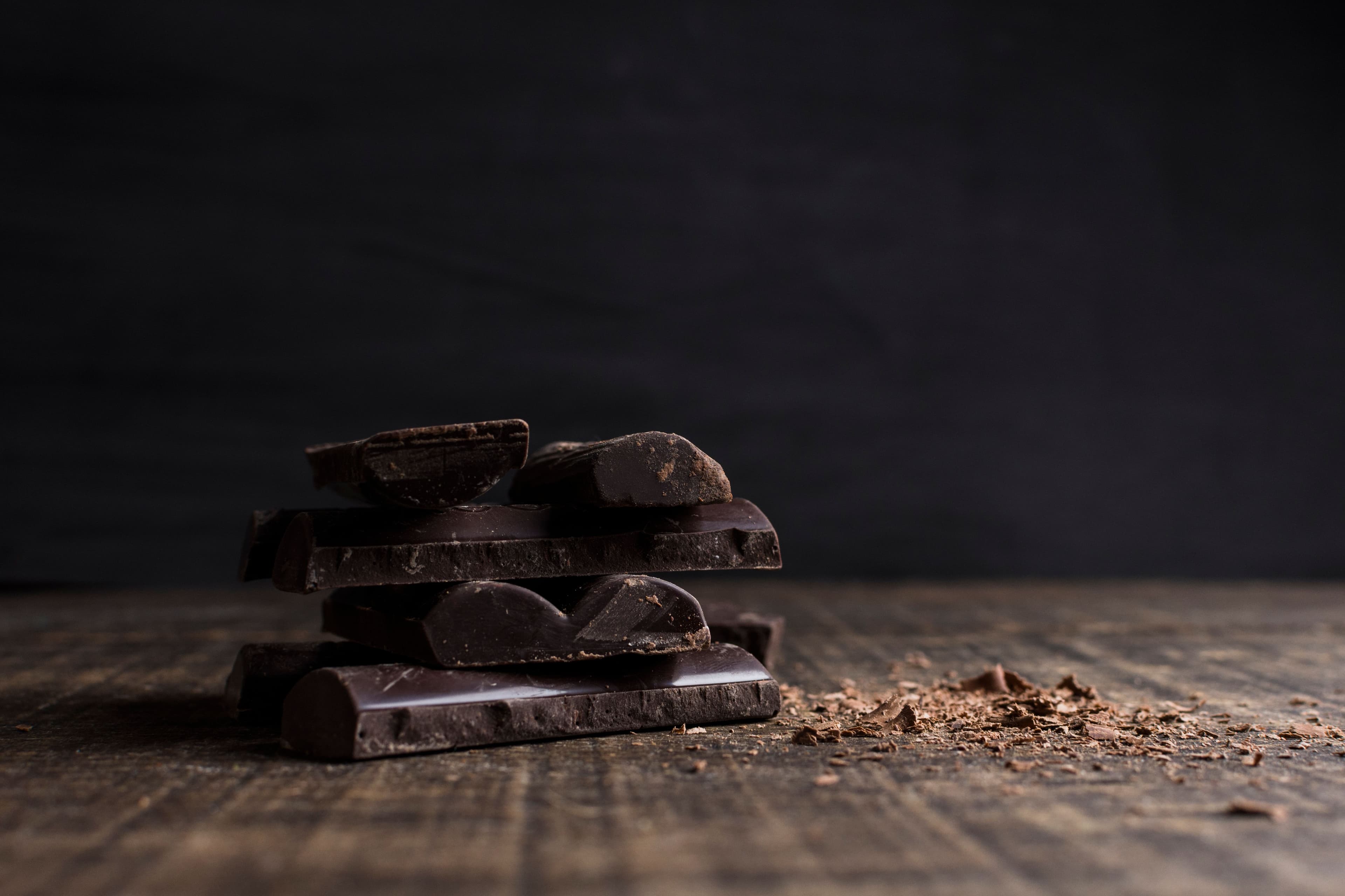A rustic stack of dark chocolate pieces with chocolate shavings on a wooden surface.