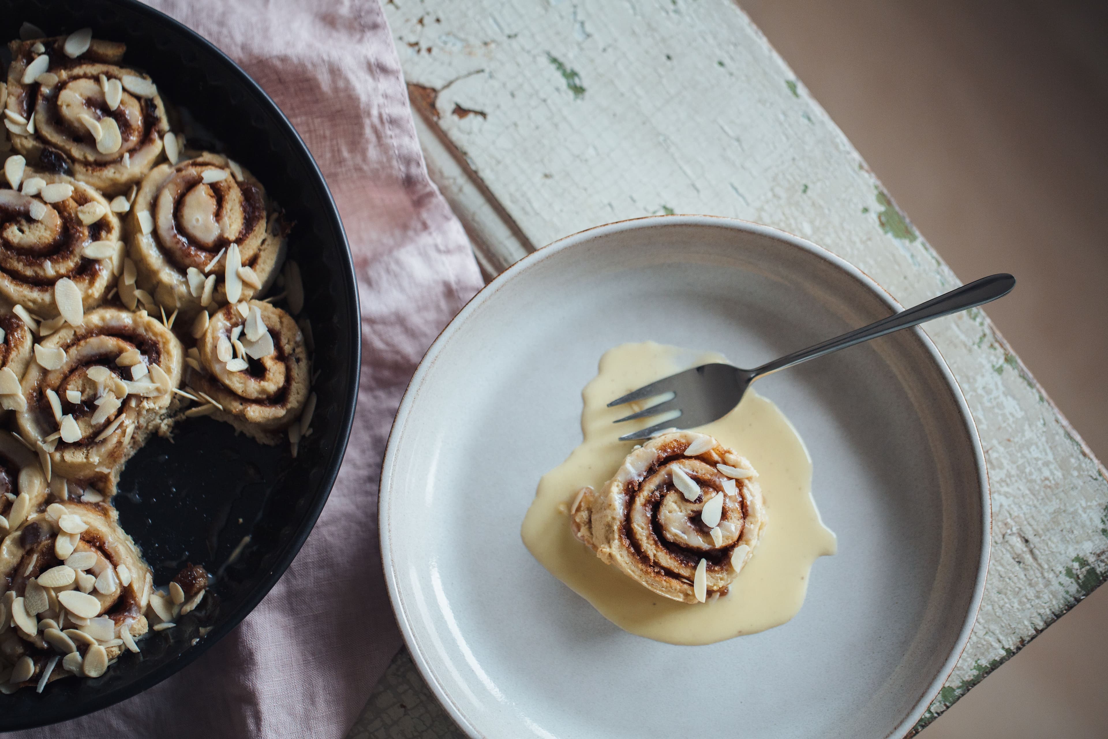 A cinnamon roll placed on a white plate beside which sits a black tray full of several cinnamon rolls.