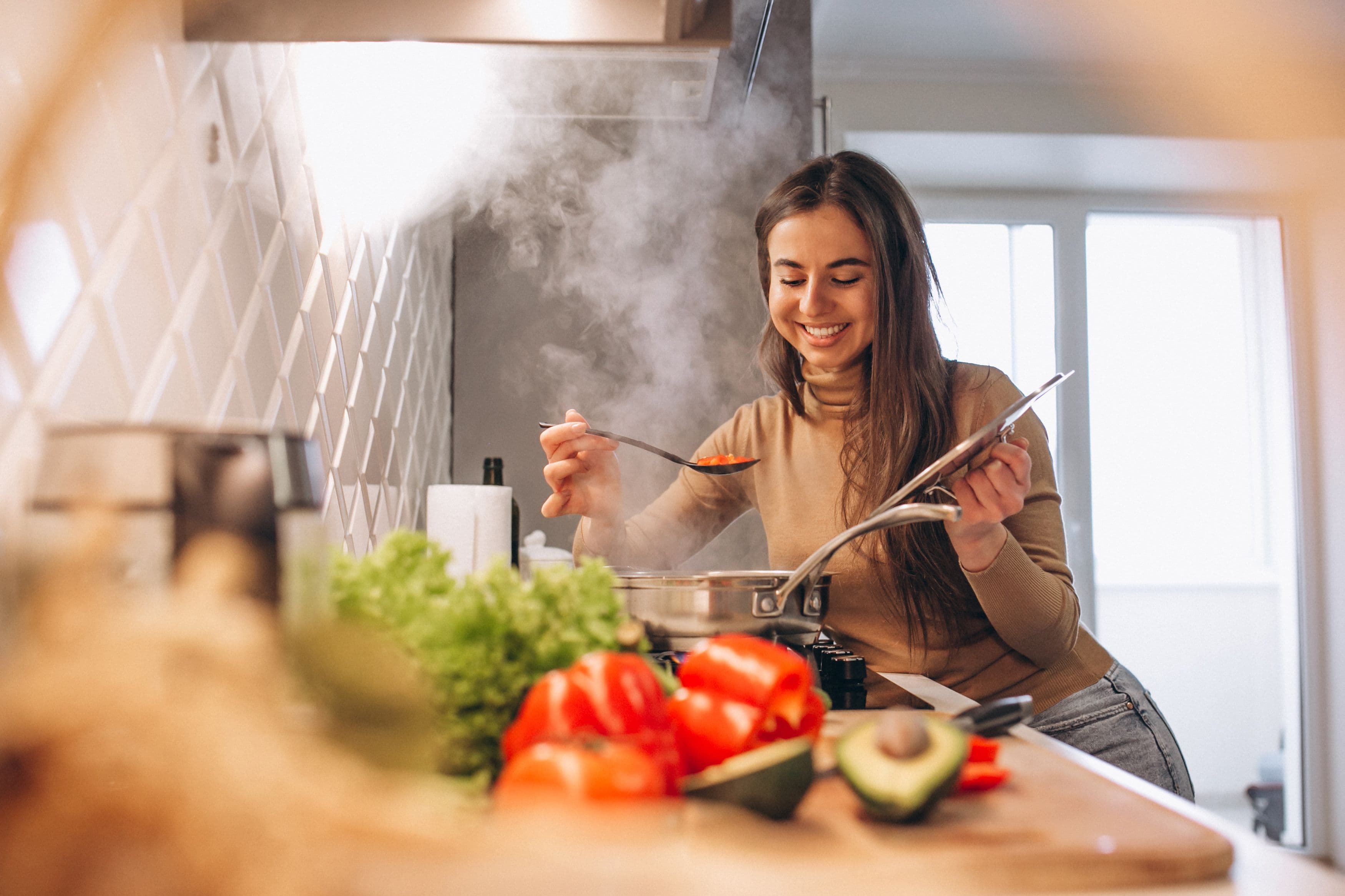 Woman smiling while tasting food from a pot in a home kitchen, surrounded by fresh vegetables