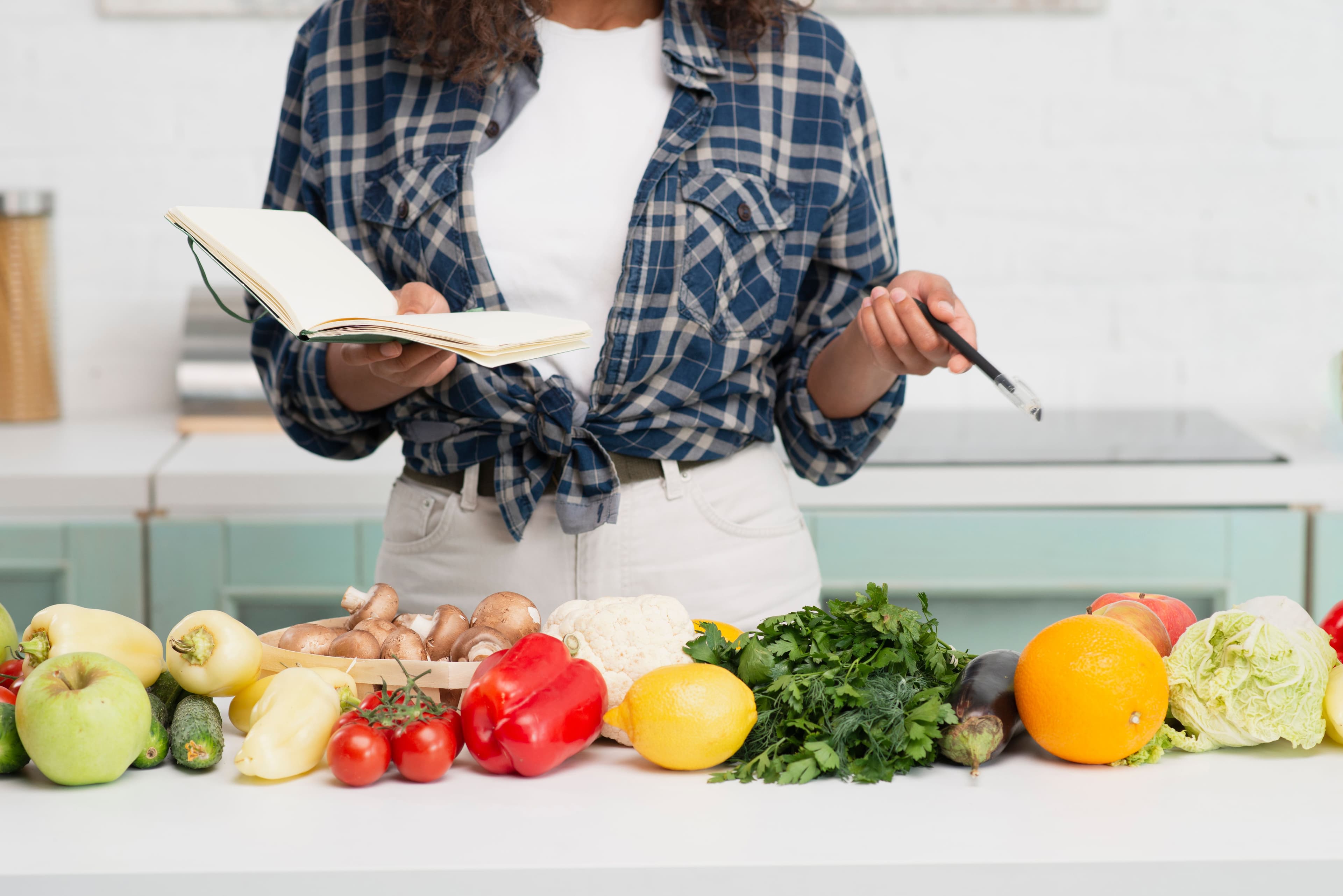 Home cook holding a notebook and pen while surrounded by fresh vegetables, planning a recipe