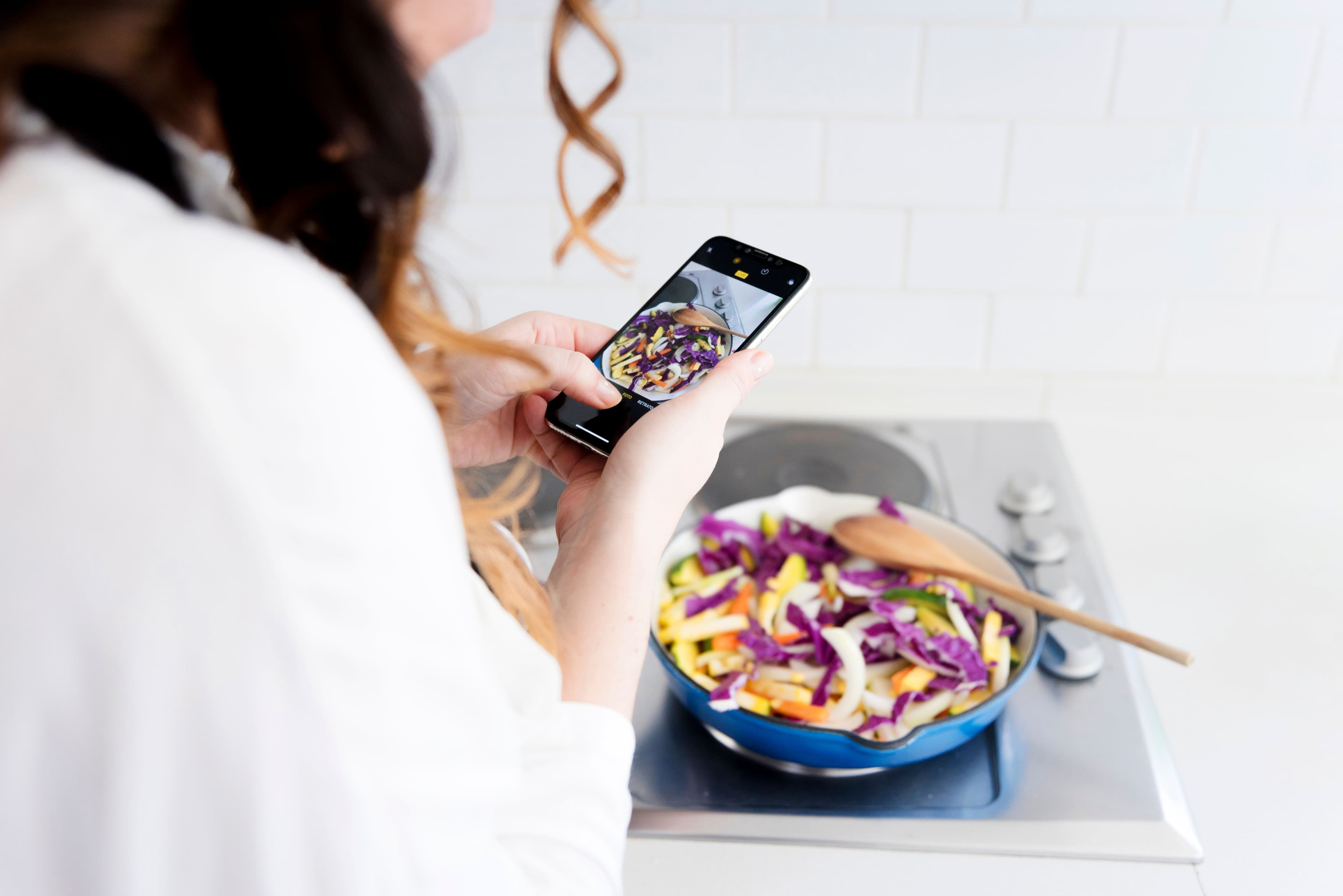 A woman capturing a photo of a colorful vegetable stir-fry in a skillet using her smartphone, showcasing food photography in a bright kitchen.