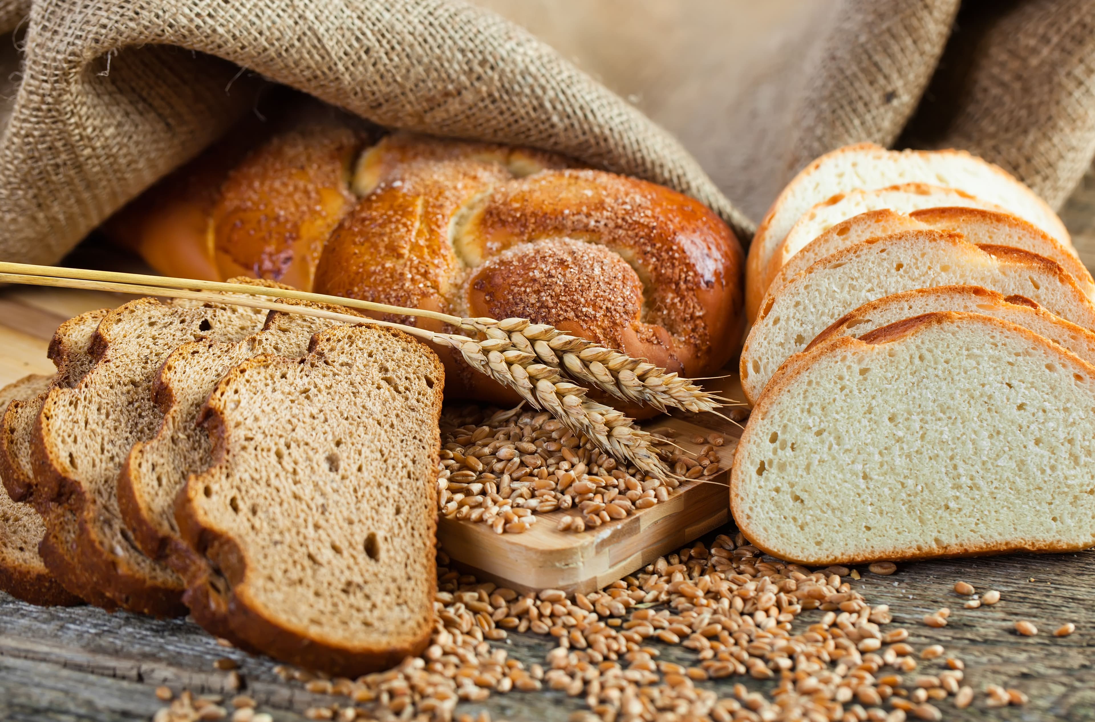 Assortment of homemade bread including braided challah, whole wheat, and white loaf slices, surrounded by wheat grains and burlap fabric—perfect for cozy weekend baking vibes.