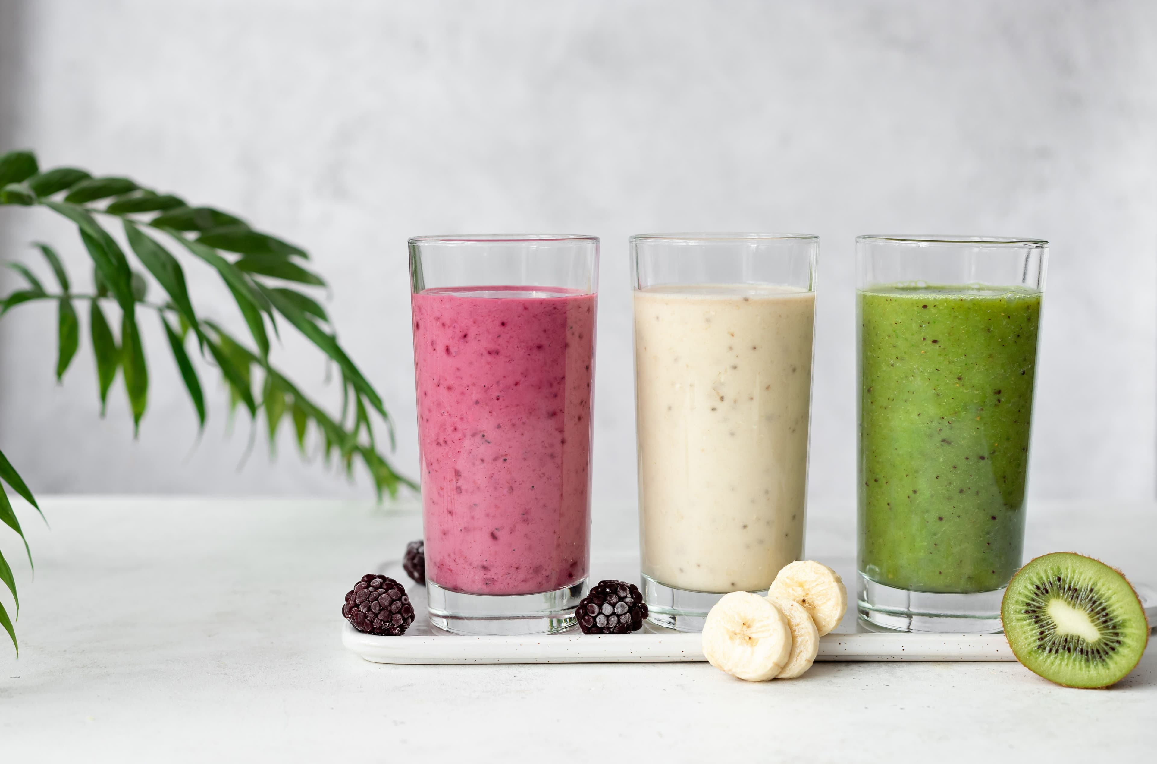 Assortment of smoothies featuring blackberry, banana, and kiwi-spinach blends on a grey background with fresh greens.