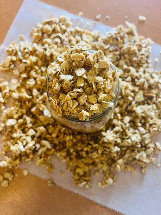 Homemade oat granola overflowing from a glass jar on parchment paper