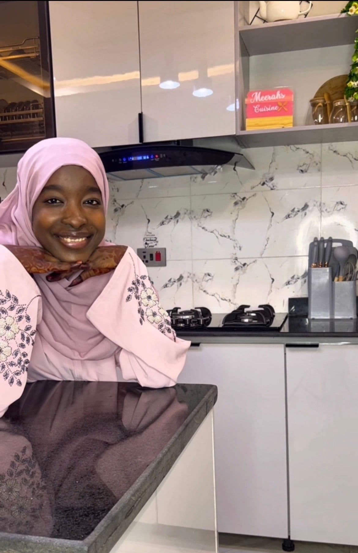 Maryam Auwal posing in her kitchen studio, wearing a light pink hijab and floral-embroidered sleeves