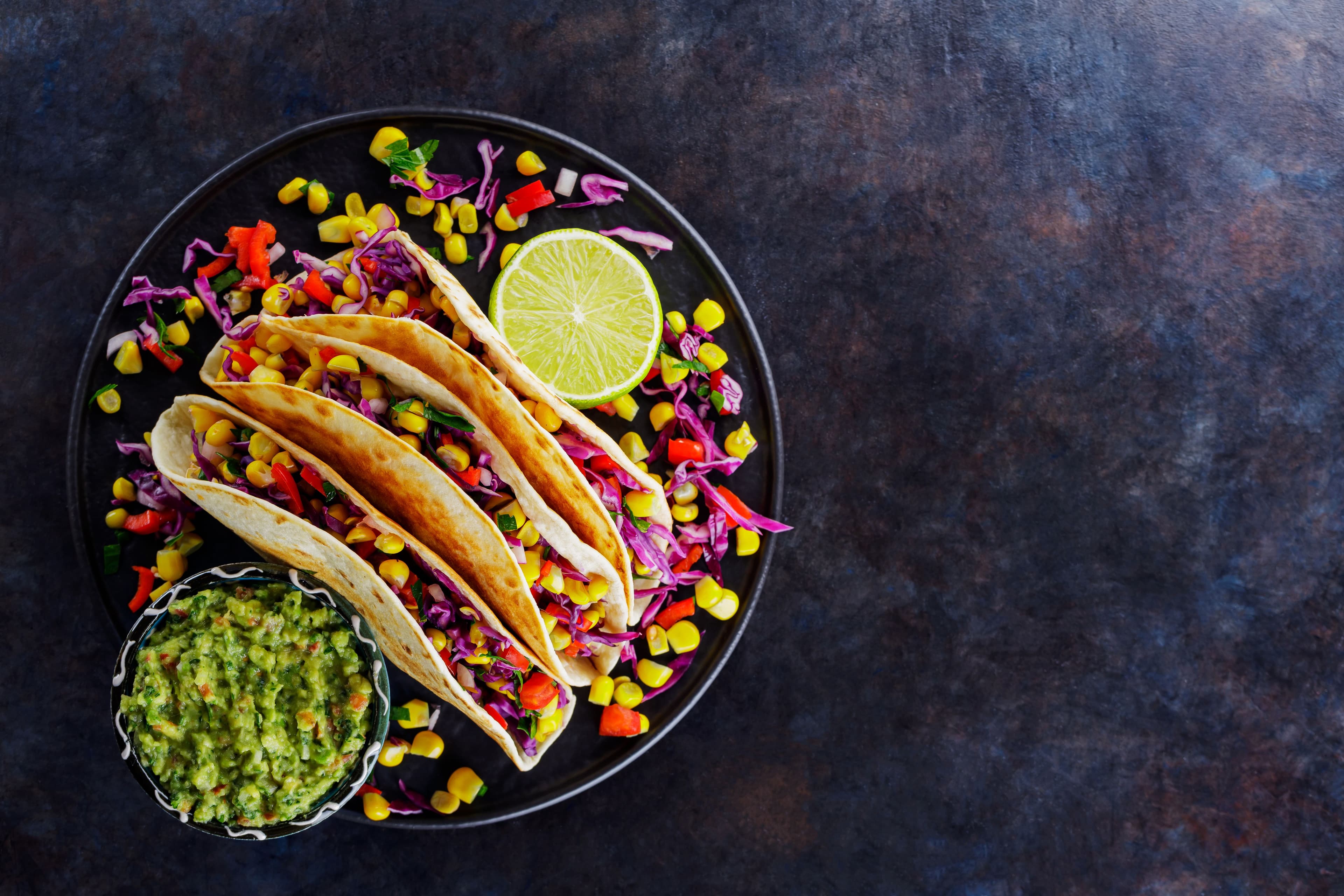 Vegetarian tacos filled with corn, red cabbage, and peppers on a dark plate, served with guacamole, the classic dish of Mexican cuisine..