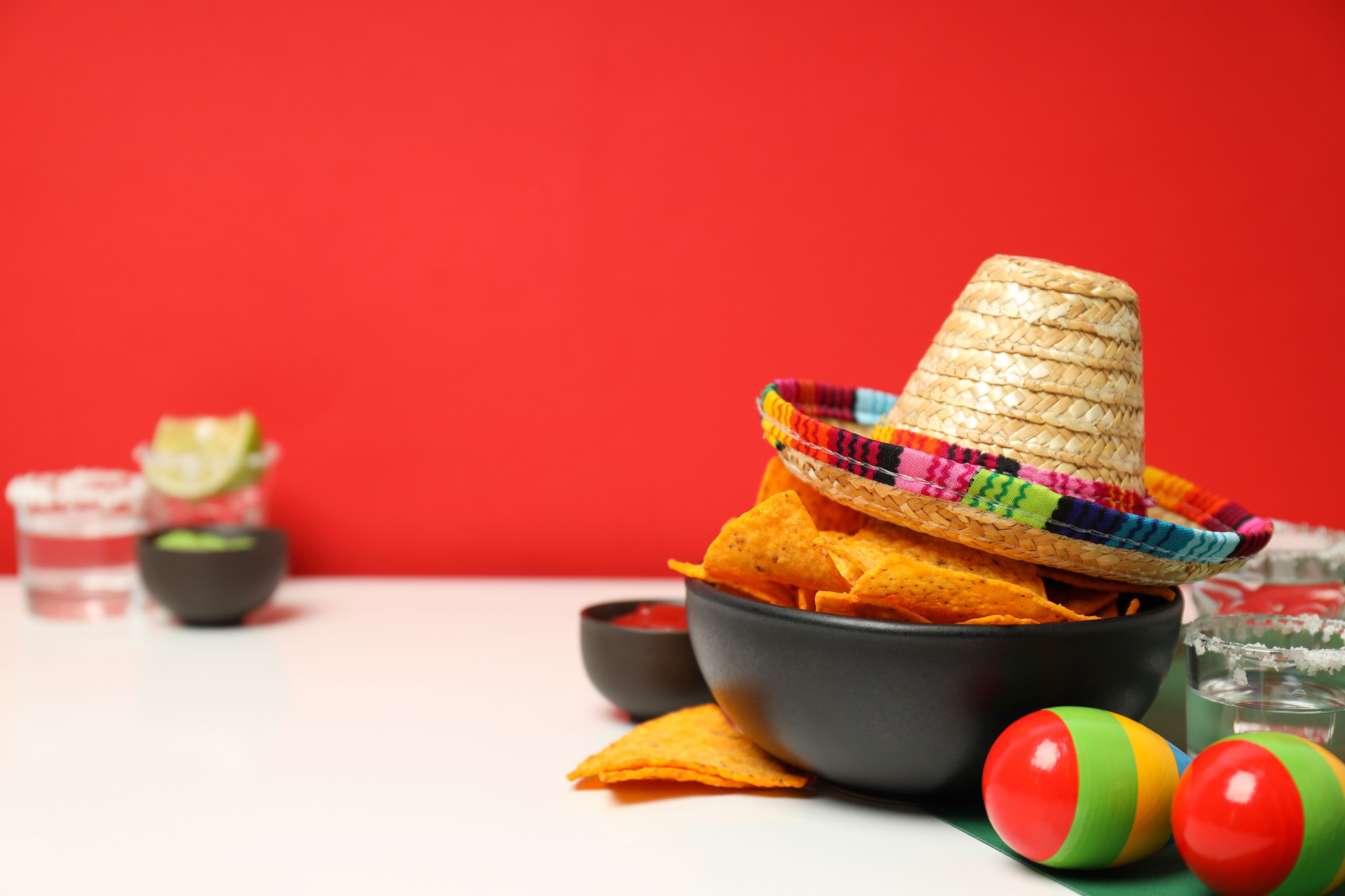 Festive Mexican cuisine setup with nachos, a colorful sombrero, and tequila shots against a bright red background.