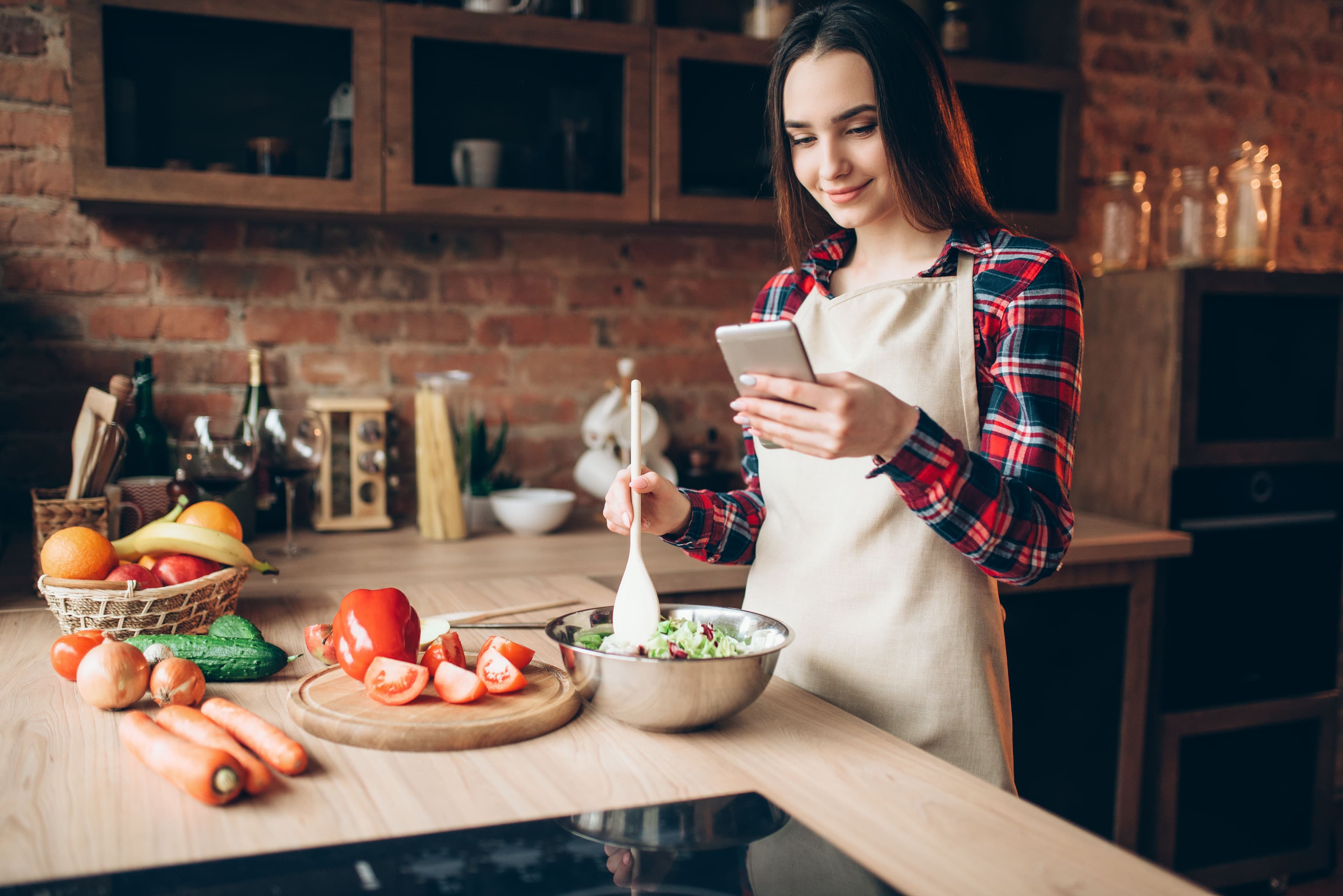 Young woman reading recipes in a modern kitchen with fresh ingredients on the counter.