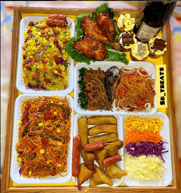 A colorful Nigerian food platter by Success Nwaokoro featuring jollof rice, fried rice, chicken wings, spring rolls, salad, and cupcakes