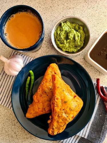 2 pieces of fried gram-flour dipped bread slices on a plate, next to a cup of tea and 2 bowls of green and brown chutneys