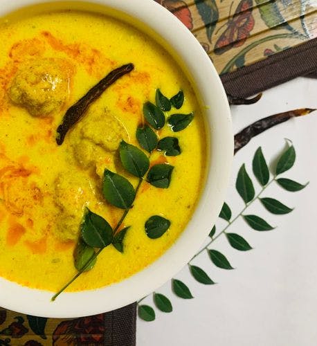 A bowl of yellow kadhi garnished with a dry red chilli and a stem of curry leaves. 