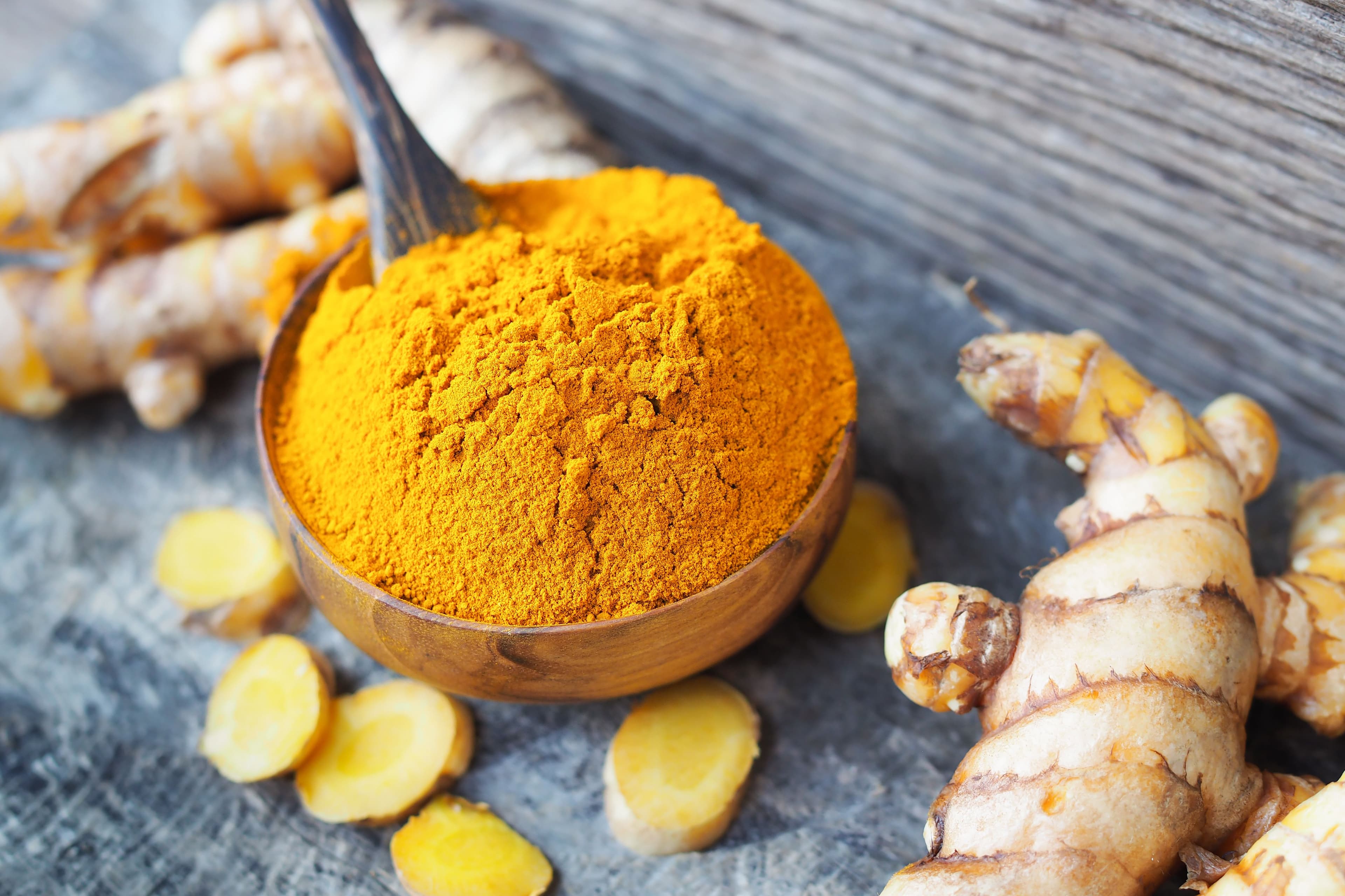 Close-up of a wooden bowl filled with bright orange turmeric powder with a wooden spoon inside, surrounded by fresh turmeric roots and sliced turmeric pieces on a rustic wooden surface.