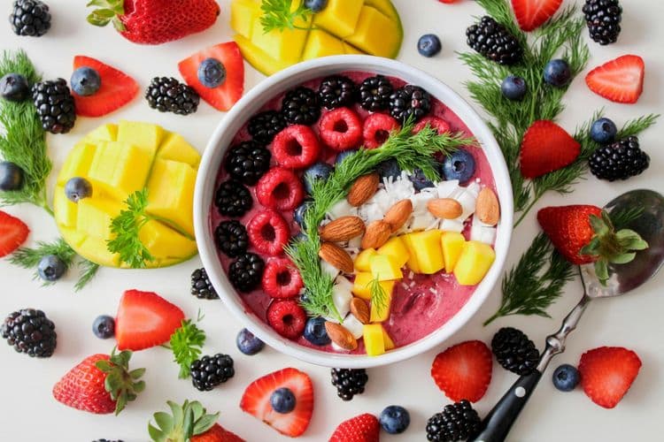 A nutritious bowl of fruit and nuts placed surrounded with several sliced fruits and a spoon.